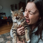 Mulher segurando um gato peludo enquanto ambos parecem espirrar ou reagir simultaneamente.